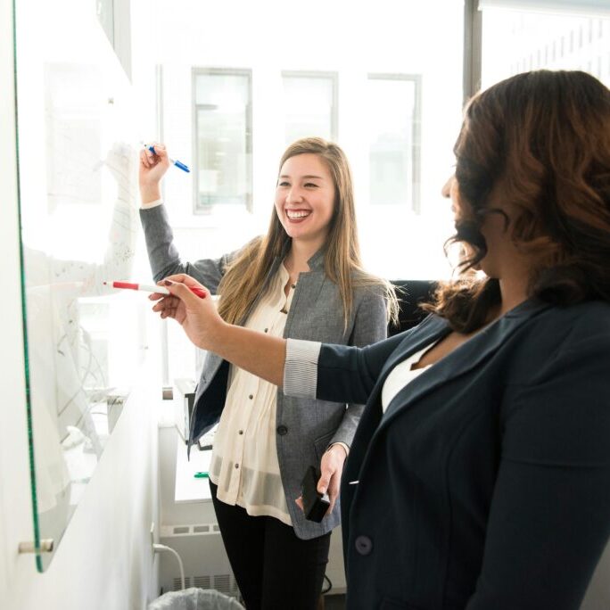 Two women in business casual attire working on a whiteboard together